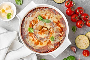 Eggplant casserole with cheese and tomato sauce in a white baking dish on a gray concrete background with ingredients for cooking