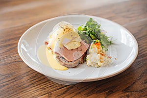 Eggs Benedict with ham, toast and mashed potato. Served with salad on a white plate on wooden table