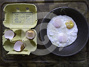 Eggs box with broken shells and fried eggs in a frying pan