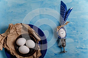 Eggs on brown paper and blue plate. Easter bunny made from egg, polka dot napkin ears in silver egg cup on wooden blue background