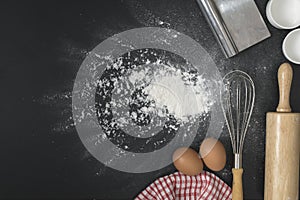 The eggs with a cup of flour and whisk on the black table spread of flour