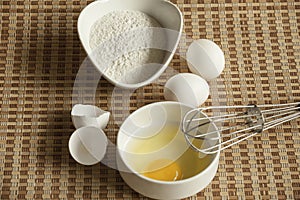 Eggs, flour and whisk on table mat.