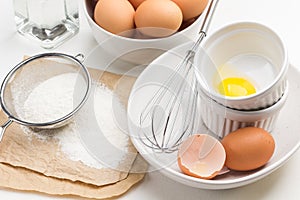 Eggshell, egg and whisk in bowl. Egg yolk in bowl. Flour and sieve on paper. Brown eggs in bowl