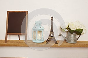 Eiffel tower statue, blue lantern, flower in metal vase and empty blackboard for copy on wooden shelves