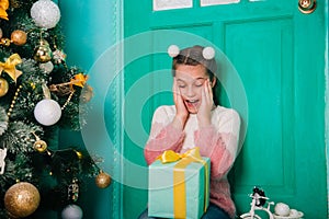 An eight-year-old girl in a pink sweater sits on the steps by the Christmas door holding a joyful present