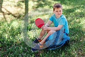 Eight-year-old sports boy in sneakers sitting on the green grass and holding a table tennis racket in his hands