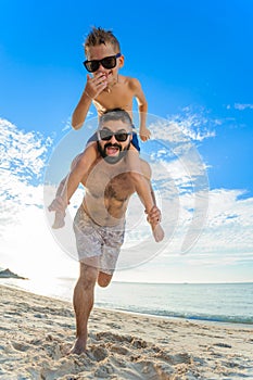 Eight years old boy sitting on dad`s shoulders. Both in swimming shorts and sunglasses, having fun on the beach. Bottom view