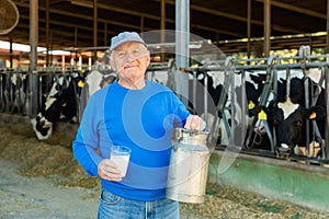 Eldelry dairy farm owner standing in cowshed with can and glass of milk