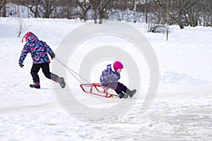 Elder girl running and pulling her young sister on a sled on the ice in snowy winter park