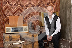 Elder grandfather in elegant clothes sitting on the old travel valise near the vintage radio, kerosene lamp and chest