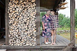 Elder grandmother and young granddaughter standing on the veranda of summer house, next to the woodpile, full-length portrait,