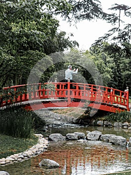 Elder man on a red bridge while taking pictures at the Japanese garden in Paris