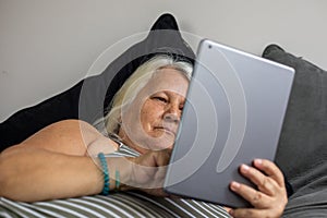 An elder obese woman laying on the bed looks at a tablet