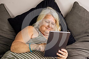 An elder obese woman laying on the bed looks at a tablet