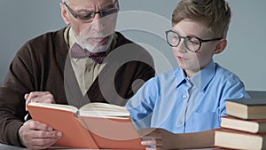 Elder teacher reading book with male pupil, explaining school subject, education