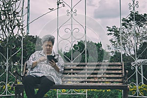 elder woman holding mobile phone while sitting on bench in garden. elderly female texting message, using app with cellphone in pa