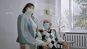 Elder woman and ill grey haired old man in protective medical masks look at the camera, male patient sitting on