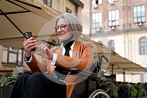 elder woman in wheelchair holding mobile phoneoutdoor. elderly female texting message, using app with cellphone in park