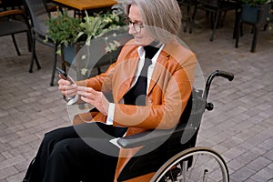 elder woman in wheelchair holding mobile phoneoutdoor. elderly female texting message, using app with cellphone in park