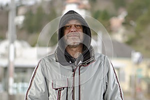 An elderly 50-55 year old hooded man with a graying beard stands confidently against a blurred suburban landscape.