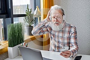 elderly 70s man browsing internet on modern laptop and listen to music using smartphone