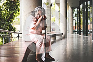 Elderly Asian female patient holding a cane Sitting sadly on a bench outside the building.