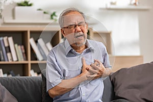 Elderly Asian male with from heart attack sitting at the couch in the living room. senior clutching his left chest from acute pain