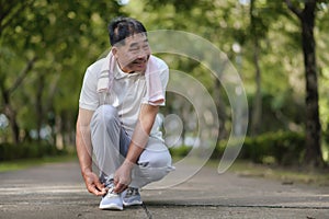 An Elderly Asian Man Is Tying Shoelaces With A Kind Face And A Gentle Smile In The Park Is Full Of Trees. Looks Around The Park