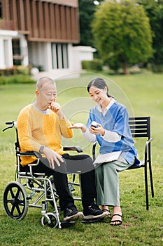 Elderly asian senior man on wheelchair with Asian careful caregiver and encourage patient, walking in garden. with care from a