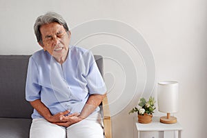 Elderly Asian woman with grey hair sits and holding her stomach in pain on the sofa, Aging society and Various illnesses of the