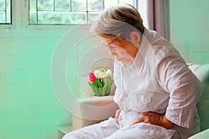 Elderly Asian woman with grey hair sits and holding her stomach in pain on the sofa, Aging society and Various illnesses of the