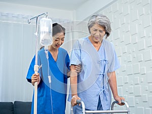 Elderly Asian woman patient trying to walk on walking frame held and carefully supported in arms by caregiver.