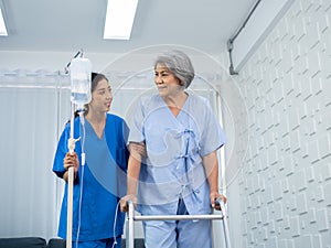 Elderly Asian woman patient trying to walk on walking frame held and carefully supported in arms by caregiver.