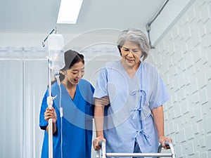 Elderly Asian woman patient trying to walk on walking frame held and carefully supported in arms by caregiver.