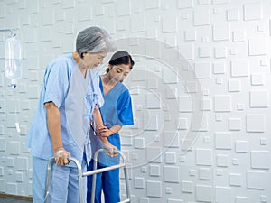 Elderly Asian woman patient trying to walk on walking frame held and carefully supported in arms.