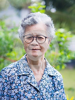 Elderly Asian woman wearing glasses, smiling and looking at the camera while standing in a garden. Concept of aged people and