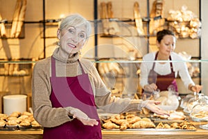 Elderly bakeshop saleswoman in maroon apron recommending baked goods