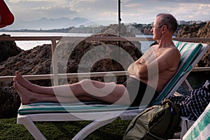 An elderly bald man with glasses lies on a sun lounger and sunbathes on the seashore in Antalya Turkey among the rocks