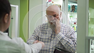 Elderly bearded man with toothache coming to the pharmacy. Young female pharmacist offering the customer pain medication