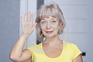An elderly beautiful woman of 60-65 years old raised her right hand in a welcoming gesture against a neutral background in the roo
