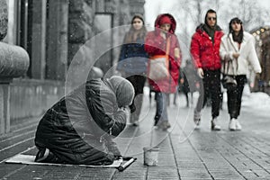 Elderly beggar woman on her knees asking for food from brightly dressed passers-by on the street