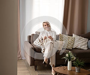 elderly blond woman sit on couch at home holding knitting needles and yarn knits clothes for loved ones