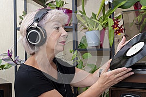 An elderly blonde woman 60-65 years old in old headphones holds a vinyl record in her hands, a record player against the backgroun