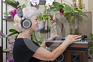 An elderly blonde woman 60-65 years old in old headphones looks at a vinyl record, a record player against the background of indoo