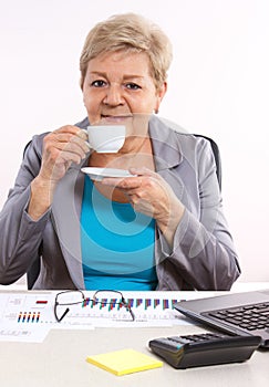 Elderly business woman drinking tea or coffee at desk in office, break at work