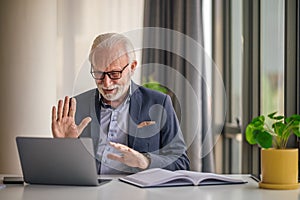 Elderly businessman greeting while video conferencing on laptop at office desk