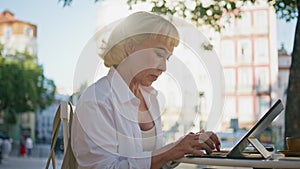 Elderly businesswoman typing laptop at sunny street cafe closeup. Serious senior