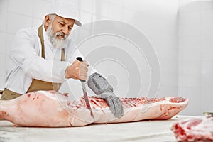 Elderly butcher in uniform cutting pork carcass with knife.
