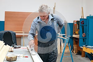 An elderly cabinetmaker in overalls and glasses paints a wooden board with a roller on a workbench in a carpentry shop.