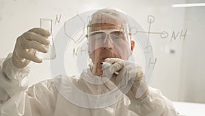 An elderly Caucasian male chemist in a protective suit holds a test tube and writes a formula on the glass.